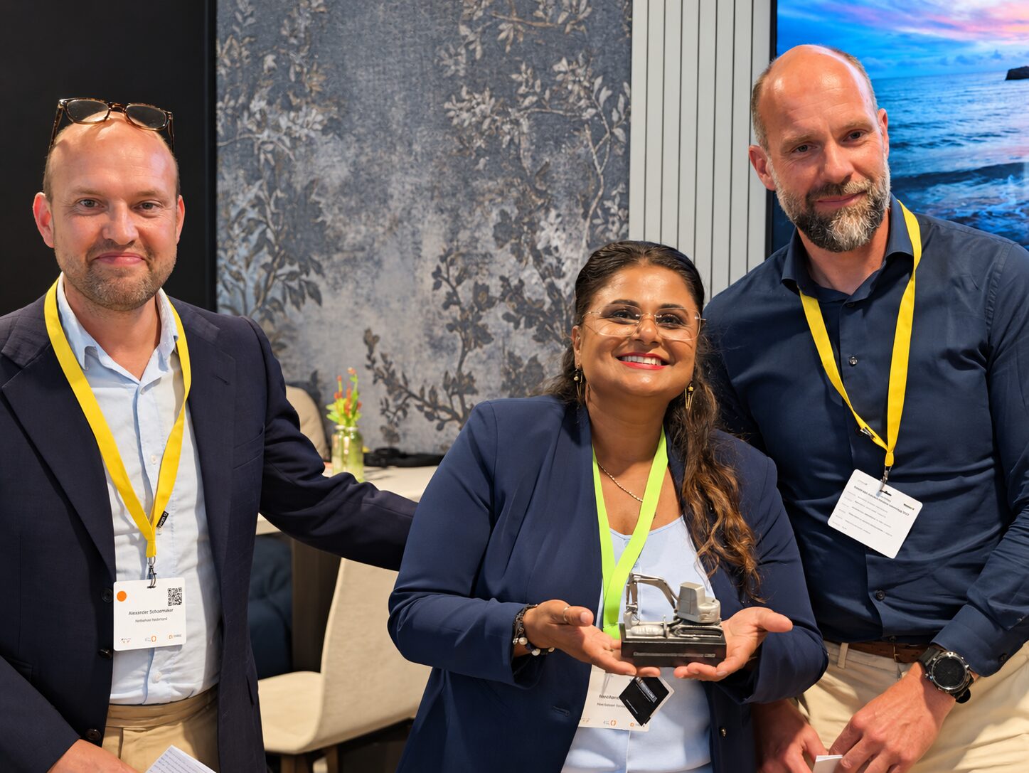 Nita Sardjoe houdt glimlachend de Graafschadepreventie Inspiratieprijs vast, een beeldje van een graafmachine, met naast haar twee mannen met congresbadges
